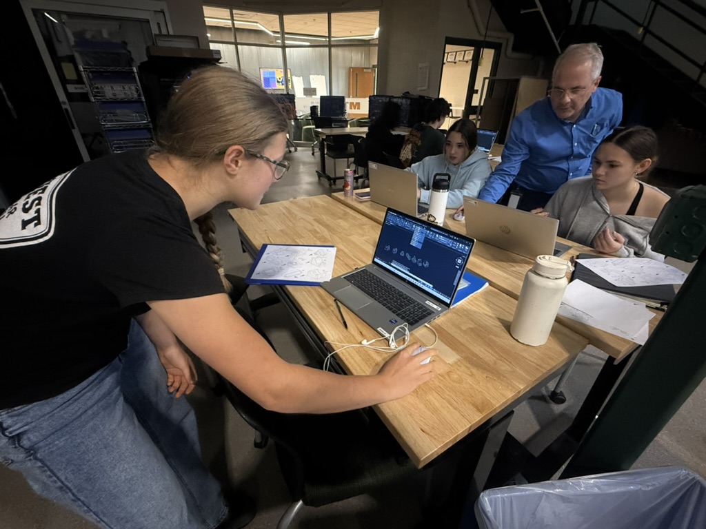 student standing at table working on laptop while another teacher and students work at another table