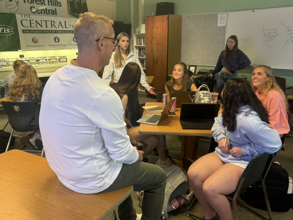Adult sitting on a desk talking to a group of students at a table
