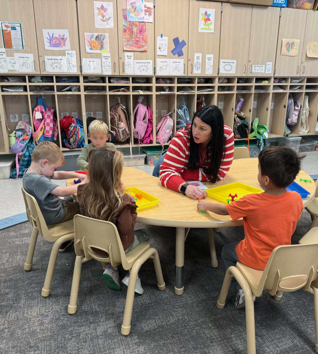 A light-skinned woman with long dark hair wearing a red and white striped sweater sits at a crescent shaped table with four small children seated around it in a pre-school classroom.