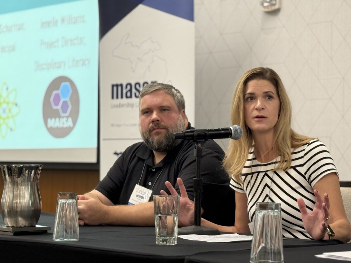 A light-complected man with dark graying hair and beard, wearing a dark short-sleeved shirt sits next to a woman light complected woman with shoulder-length blond hair who is wearing a blue and white striped, short-sleeved shirt who is speaking into a microphone at a speaker panel table.
