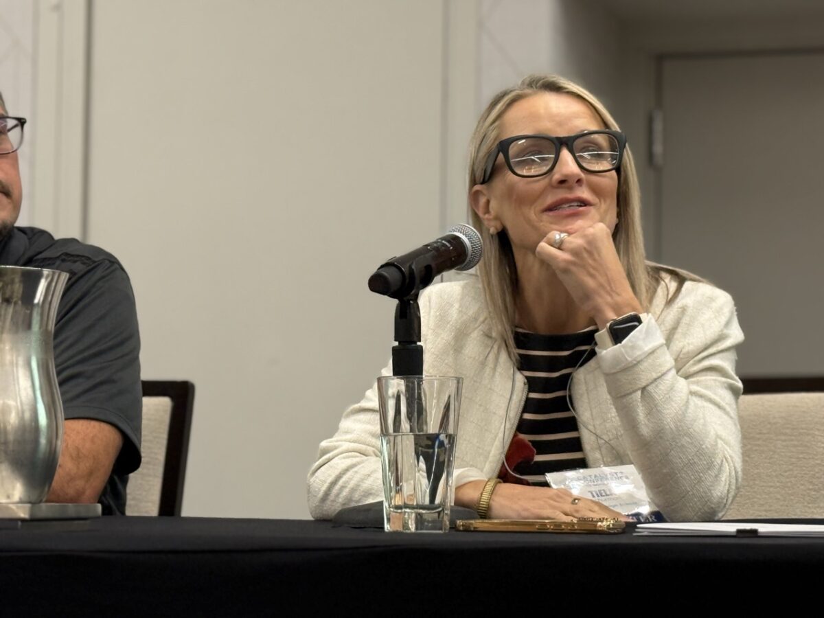 A light-complected woman with blond hair, wearing black, thick-framed glasses, a black and white striped top under a white jacket and a bold watch speaks into a microphone while sitting at a speaker panel table.