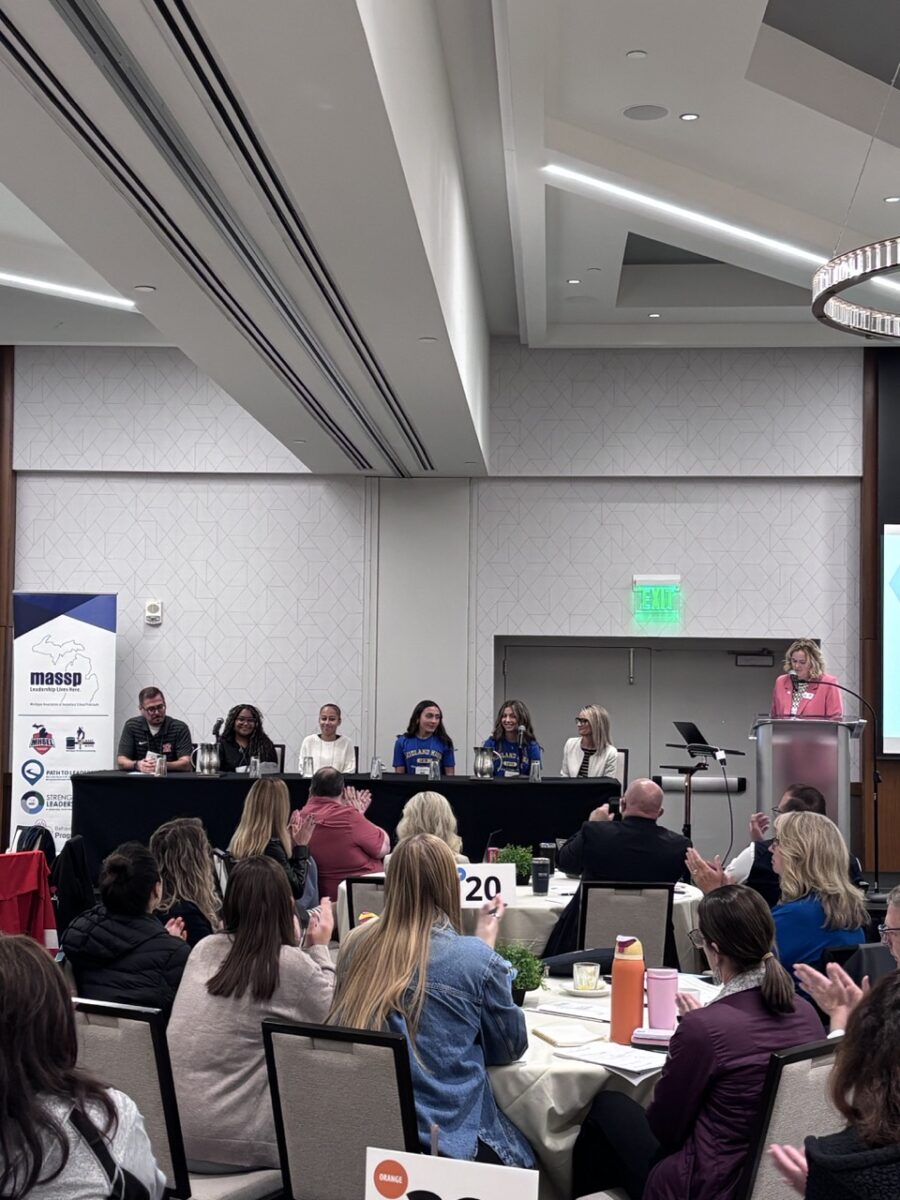 From a distance, beyond multiple tables of people meeting at a conference, there is a panel with guest speakers, a mix of students and educators.