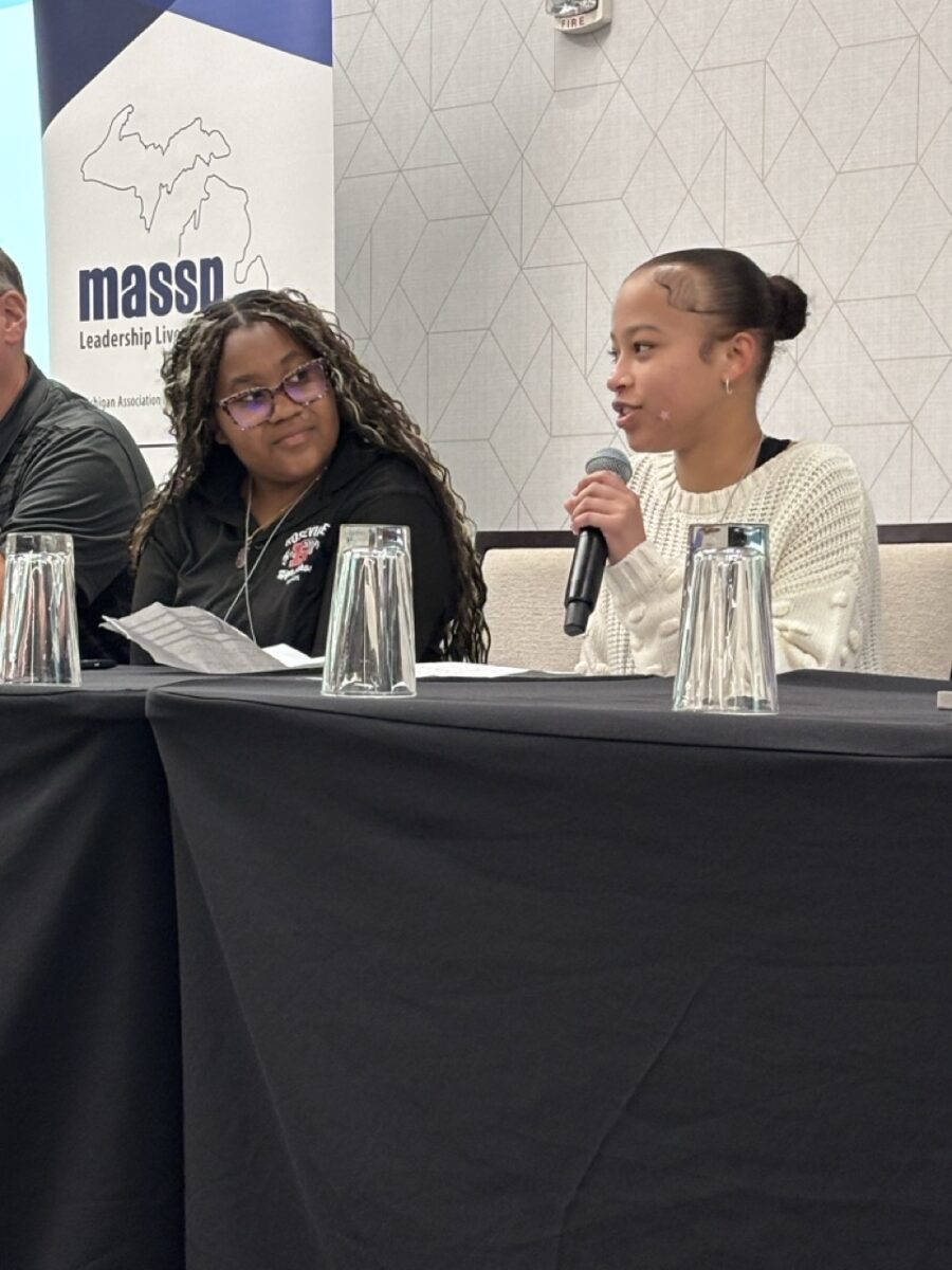 A medium-complected young woman with her dark hair pulled back into a bun, wearing a cream-colored sweater speaks into a microphone she is holding while sitting at a speaker panel table. To her right, a dark-complected young woman with long dark, curly hair wearing glasses watches her speak.