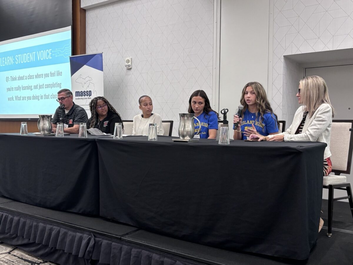 A group of two adult educators, one man and one woman, sit at a panel of speakers with four female students at a long table with a black cover over it.