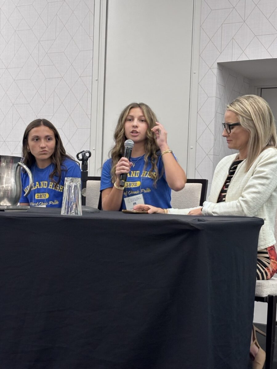 A light-complected young woman with long wavy, light-brown hair wearing a blue T-shirt with yellow writing on it who is holding and speaking into a microphone sits between a fellow student and her principal at a speaker panel table.