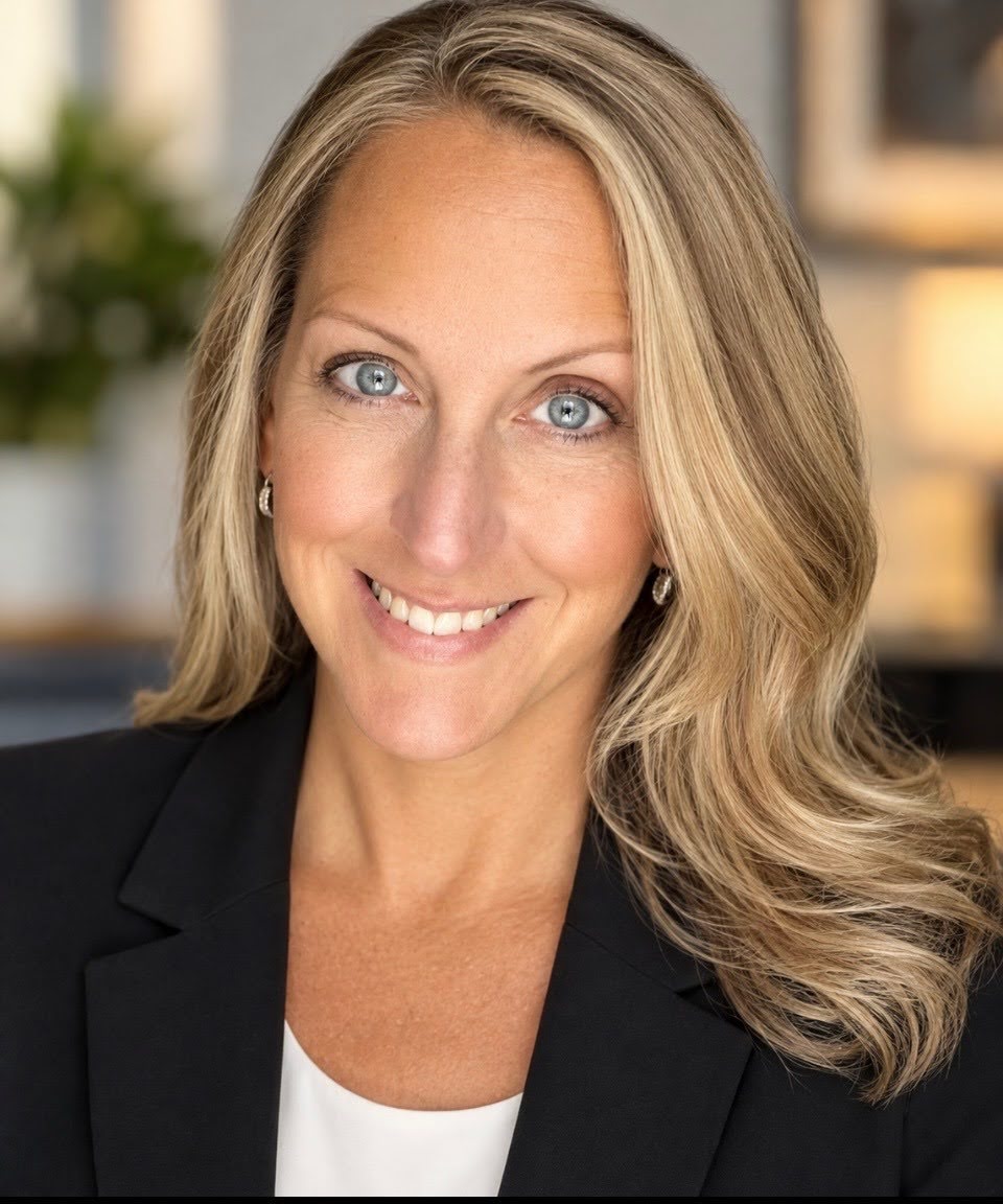 A blond-haired woman with blue eyes wearing a white T-shirt and a dark blazer smiles. This is a portrait image.
