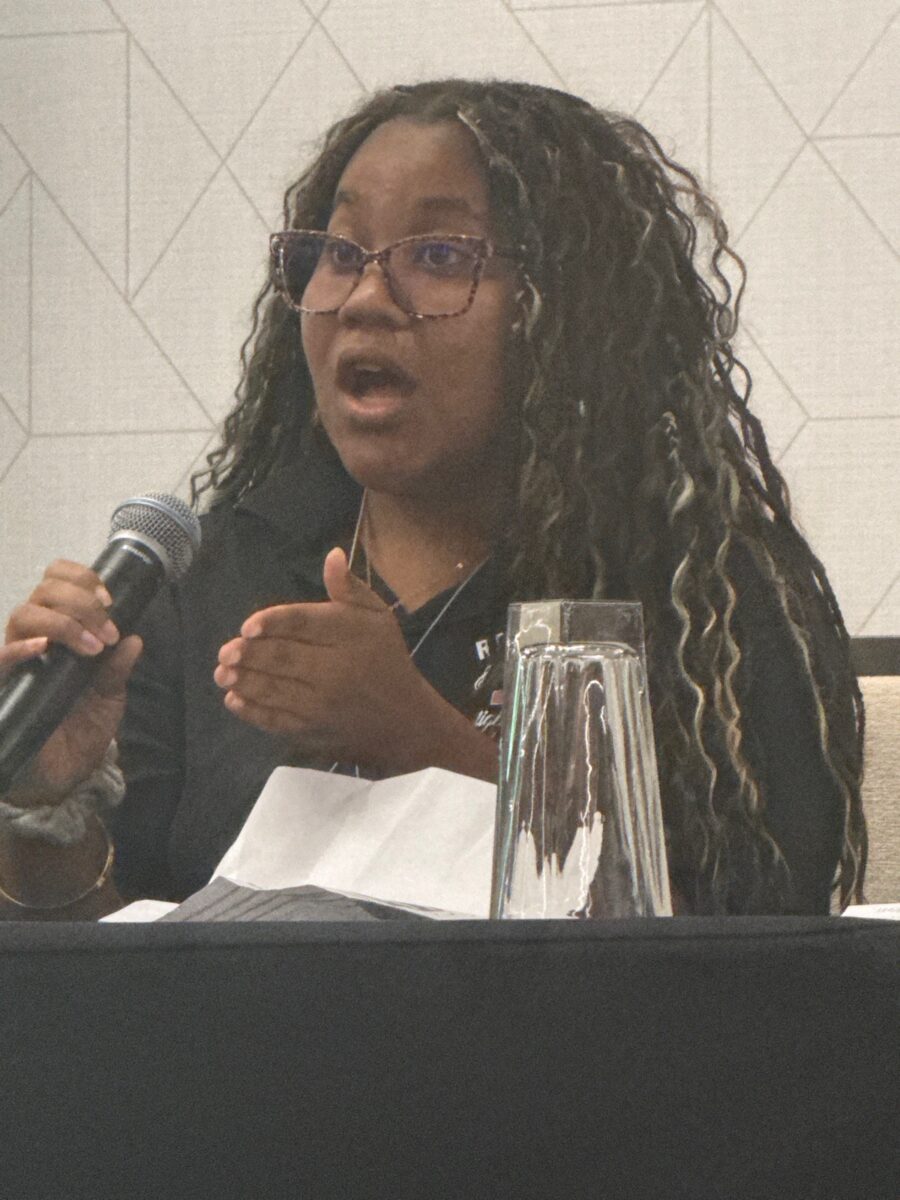 A dark-complected, young woman with long curly hair wearing glasses and a dark long-sleeved shirt gestures with her left hand while holding a microphone in her right hand and speaking into it. She is seated at a panel speaker table.
